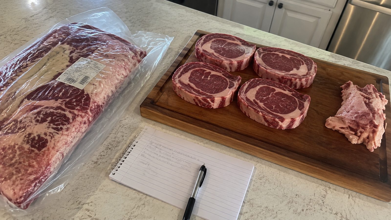 A whole cryovac ribeye primal beside four cut steaks, a pile of trim, and a notebook.