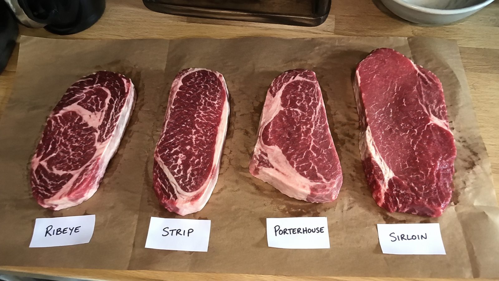 Four different raw beef steaks laid out on butcher paper with handwritten labels.