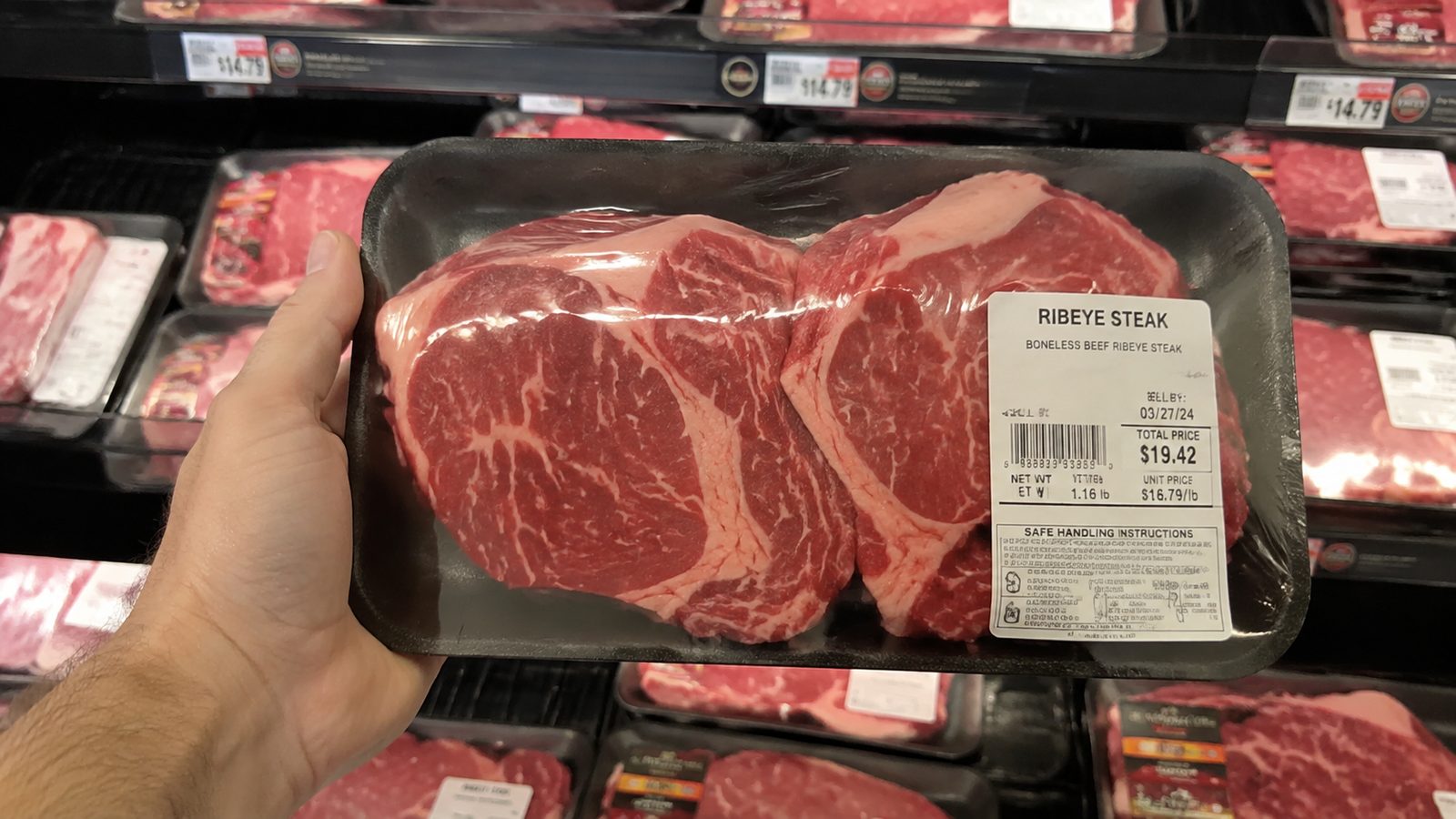 A hand lifting a tray of two ribeye steaks off the shelf of a supermarket meat case.