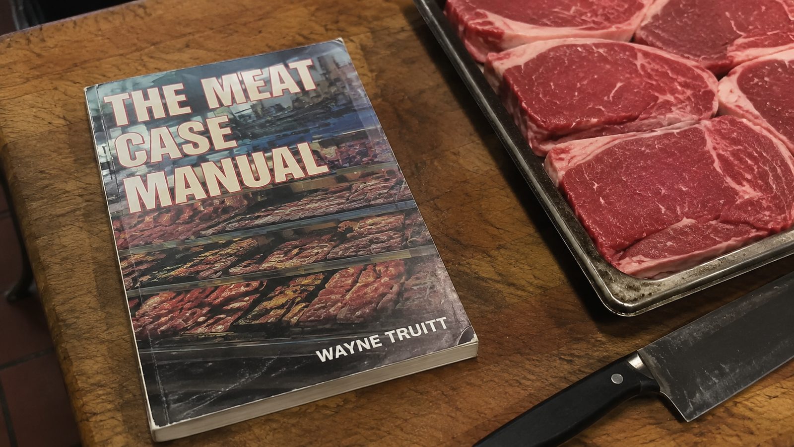 The Meat Case Manual lying on a butcher block next to a tray of steaks and a knife.